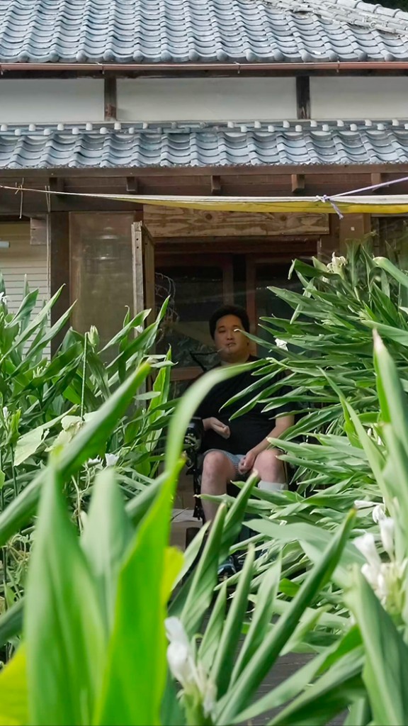 緑に囲まれた縁側でくつろぐ男性と日本家屋